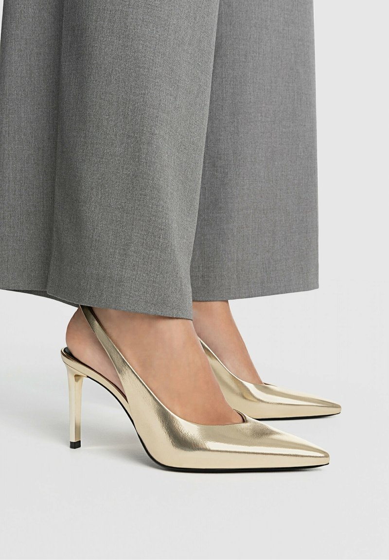 Escarpins slingback métalliques dorés avec bouts pointus et talons aiguilles fins, associés à un pantalon gris à jambes larges, mettant en valeur une texture lisse.