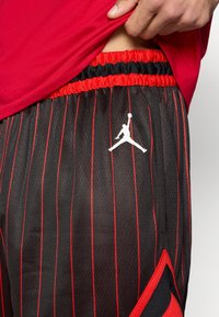 Μαύρο σορτς μπάσκετ με κόκκινες λεπτές ρίγες, ελαστική μέση και λευκό λογότυπο Jordan. Διακοσμημένο με κόκκινη τριγωνική λεπτομέρει α στο πλάι.