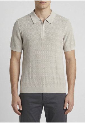 Muškarac u bež teksturiranoj polo majici kratkih rukava sa zatvaračem na ovratniku i tamnosivim hlačama, stoji ispred jednostavne pozadine.