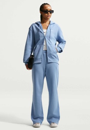 Kobieta ubrana w jasnoniebieski dres Nike z bluzą z kapturem na zamek, białą koszulkę na ramiączkach, trampki, czarne okulary przeciwsłoneczne i trzyma czarną torbę.
