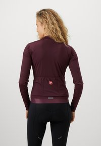 Bordeauxfarbendes Langarm-Radsport-Trikot mit einem runden Logo auf dem Rücken, entworfen mit einer enganliegenden Passform und einer strukturierten Oberfläche. Schwarze Radsportshorts sind enthalten.