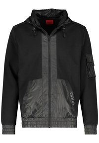 Chaqueta negra con cremallera y capucha, que presenta un panel frontal texturizado en gris oscuro, puños elásticos y un bolsillo lateral con solapa de velcro.