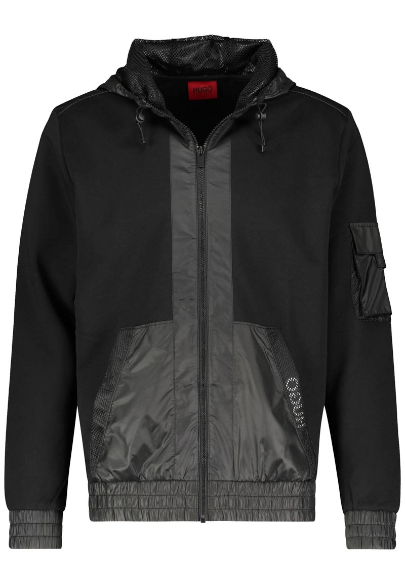 Chaqueta negra con cremallera y capucha, que presenta un panel frontal texturizado en gris oscuro, puños elásticos y un bolsillo lateral con solapa de velcro.