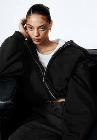 Crna prevelika majica s kapuljačom i patentnim zatvaračem, uparena sa sivim hlačama visokog struka. Glatki materijal s opuštenim krojem.