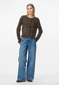 Brun stickad kofta med volangkant och knappfram svängning, ihop med vida blå jeans. Modellen står mot en vit bakgrund.