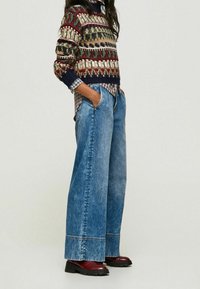 Pull en tricot avec motifs géométriques multicolores, associé à un pantalon large en denim bleu clair et des bottines rouge foncé. Détails texturés visibles.