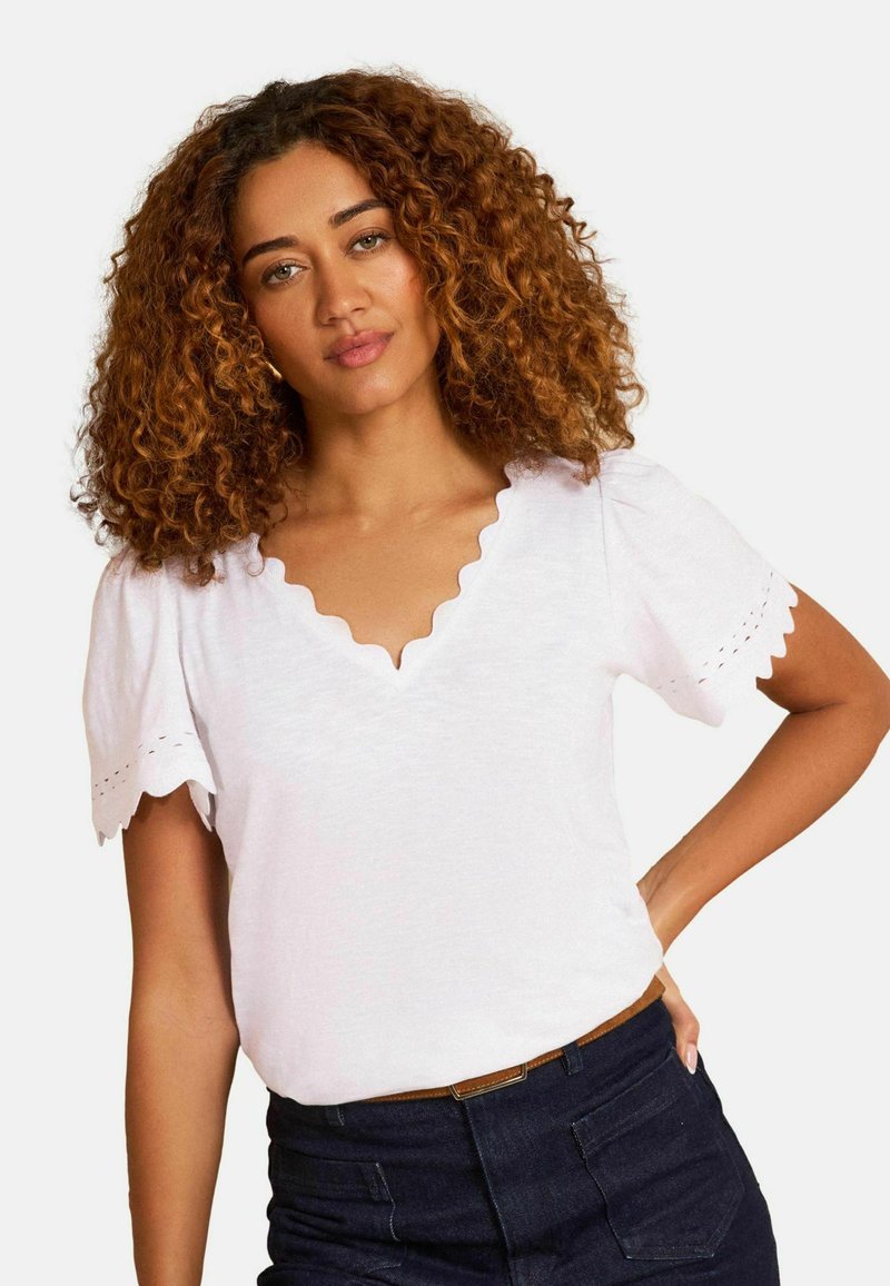 Mujer con cabello rizado, vestida con una blusa blanca con escote en V y mangas cortas con borde festoneado, y pantalones oscuros de talle alto, posando con la mano en la cadera.