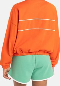 Felpa arancione a maniche lunghe con strisce decorative bianche sul retro, dotata di orlo arricciato. Abbinata a pantaloni corti verdi bordati di bianco.