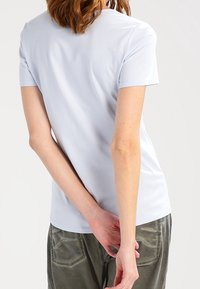 Svetle modré tričko s krátkým rukávem, přiléhavý design, hladká textura, kulatý výstřih, oblečeno s zelenými kalhotami, viděno ze zad.