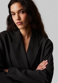 Blazer nero con un design strutturato, dotato di ampi revers e una vestibilità rilassata. Tessuto testurizzato con un'estetica pulita e minimalista.