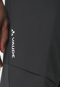 Grijze outdoorbroek met een gladde textuur, voorzien van een wit "Vaude" logo aan de zijkant en gestikte details langs de naden.