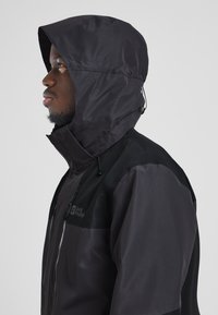 Giacca impermeabile nera con cappuccio regolabile, dotata di zip frontale, cordino regolabile e logo sul petto, texture opaca e design elegante.