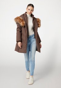 Rudas paminkštintas parkas su dirbtinio kailio akcentais ant pečių, su užtrauktuku ir šoninėmis kišenėmis, derinamas su šviesiai mėlynomis džinsais ir baltais sportbačiais.