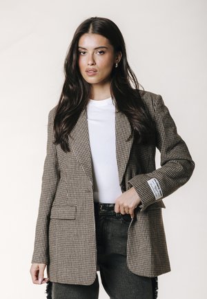 Mujer con una chaqueta marrón de pata de gallo sobre una camisa blanca y vaqueros negros, con cabello largo y oscuro y expresión neutral.