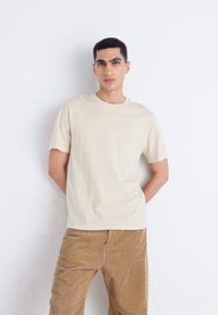 Beige katoenen t-shirt met een ronde halslijn en korte mouwen. Voorzien van een subtiele merkl logo in lichtblauw. Gecombineerd met een bruine corduroy broek.
