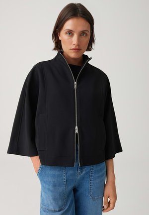 Femme portant une veste noire zippée à manches larges et un jean bleu, debout devant un fond clair uni.