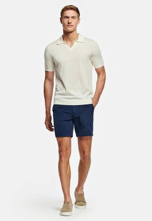 Man loopt, draagt een crèmekleurige korte mouw gebreide polo, marineblauwe korte broek en beige instapschoenen tegen een witte achtergrond.