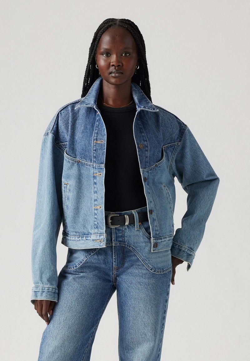 Giacca di denim con un lavaggio blu bicolore, design oversize, chiusura frontale con bottoni e tasche sul petto, abbinata a jeans in denim a vita alta.