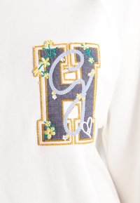 Wit kledingstuk met een grote geborduurde patch met een donkerblauwe "G" en "H", geaccentueerd met gele bloemmotieven en een hartdetail.
