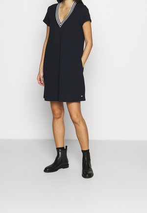 Woman wearing a short-sleeve black dress with a striped V-neckline and black ankle boots, standing with one hand in a pocket.