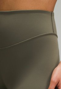 Visoke športne legice v olivno zeleni barvi, izdelane iz gladkega, raztegljivega materiala s ploskimi šivi za dodatno udobje in vzdržljivost.