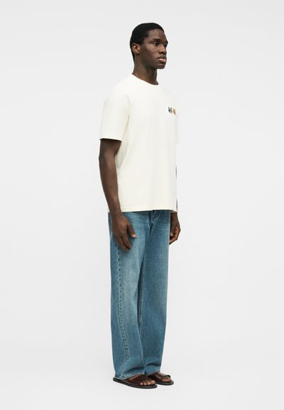 Άνδρας που στέκεται, φοράει λευκό κοντομάνικο μπλουζάκι με μικρά σχέδια στο στήθος, άνετο μπλε τζιν και καφέ σανδάλια σε λευκό φόντο.