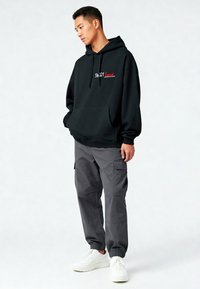 Felpa nera con cappuccio e scritta rossa "SWEET", abbinata a pantaloni cargo grigi e sneakers bianche. Vestibilità rilassata con tasca frontale.