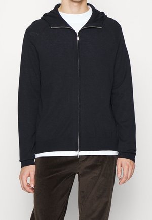 Homme portant un sweat à capuche noir zippé sur une chemise blanche et un pantalon en velours côtelé marron, debout devant un fond blanc.