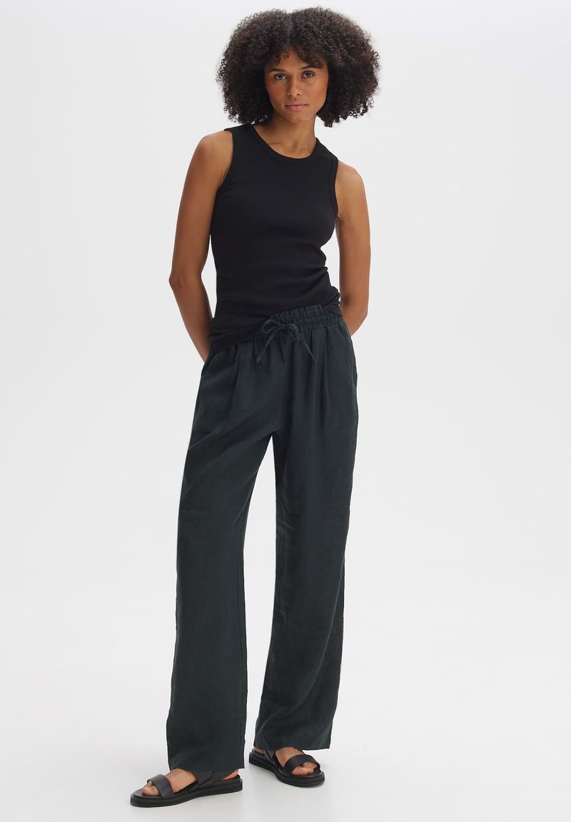 Zwarte mouwloze top gecombineerd met donkergroene, losse, met een trekkoord verstelbare broek. Het model draagt platte sandalen en heeft krullend haar.