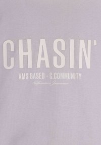 Felpa di un delicato viola chiaro con grande testo bianco che recita "CHASIN'" e un testo più piccolo sotto che dice "AMS BASED - C.COMMUNITY" in un carattere a contrasto.