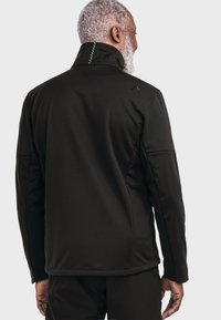 Black soft-shell jacket with a high collar, featuring green stitching on the back and a logo on the shoulder. Smooth texture and fitted design.