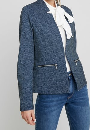Frau trägt einen strukturierten blauen Blazer mit Reißverschlusstaschen, weiße Bluse mit Schleife und blaue Jeans vor einem schlichten Hintergrund.