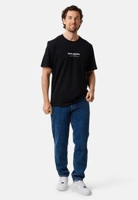 Zwarte katoenen t-shirt met witte tekst "Slade Studios" en grafisch ontwerp, gecombineerd met blauwe spijkerbroek en witte sneakers.