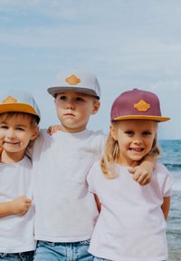 Tres niños llevan gorras diferentes: una gris con naranja, otra azul claro con naranja, y otra marrón con amarillo. Están vestidos con camisetas blancas informales y vaqueros.
