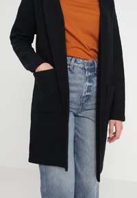 Musta pitkä neuletakki rennolla istuvuudella, valmistettu teksturoidusta kankaasta, jossa on kaksi lantiotaskua. Päällä ruosteenvärinen paita ja farkut.