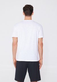 Camiseta blanca de manga corta hecha de tejido ligero, con cuello redondo y un diseño liso, llevada con pantalones cortos negros.