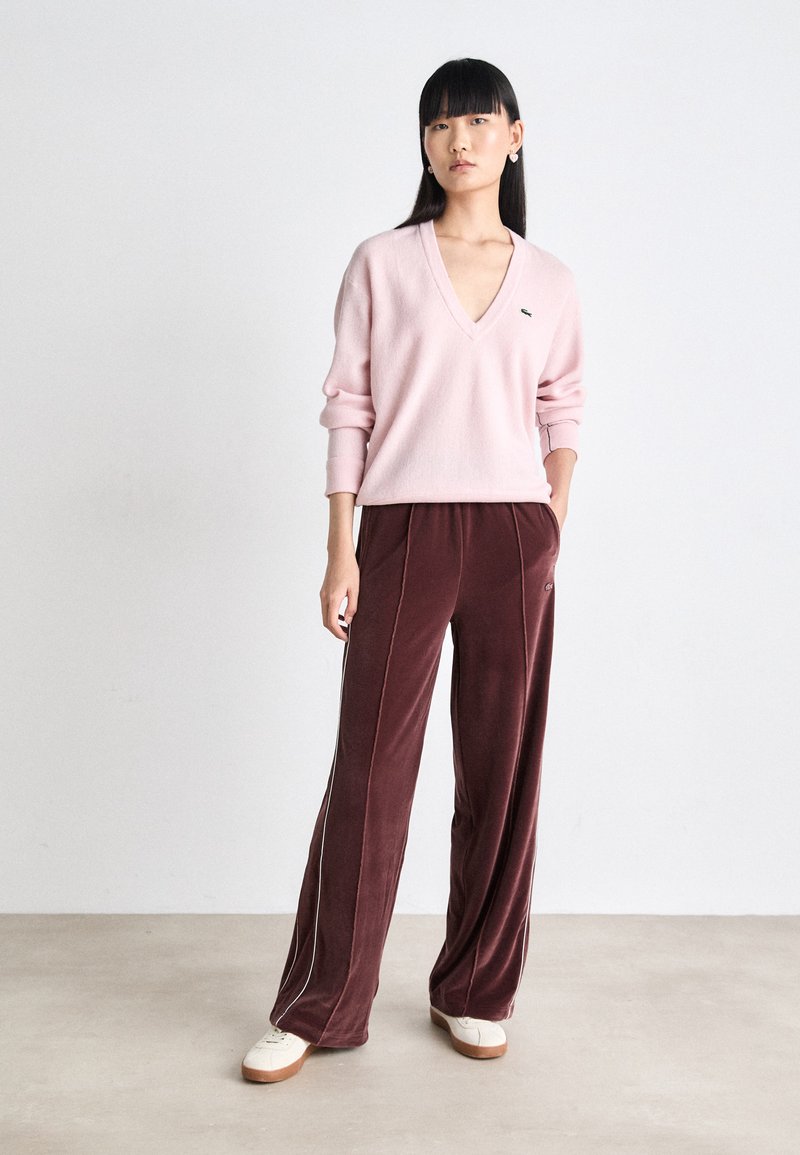 Pull à col en V rose clair en tricot doux, associé à un pantalon en velours bordeaux avec des rayures latérales et un logo assorti, le tout complété par des baskets beiges.