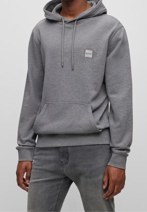 Homme portant un sweat à capuche BOSS gris avec cordons et poche avant, associé à un jean gris sur fond uni.