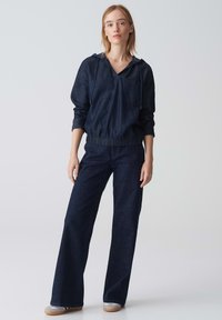 Outfit in denim scuro composto da un top con cappuccio, polsini elastici e tasca frontale, abbinato a pantaloni a gamba larga, il tutto in un uniforme colore blu scuro.