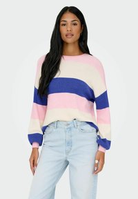 Femme aux longs cheveux noirs portant un pull rayé rose, bleu et blanc, ainsi qu'un jean bleu clair, debout devant un fond uni.