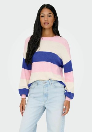 Femme aux longs cheveux noirs portant un pull rayé rose, bleu et blanc, ainsi qu'un jean bleu clair, debout devant un fond uni.