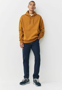 Sudadera con capucha en ámbar, con bolsillo frontal, combinada con jeans azul oscuro y zapatillas negras con detalles grises. Textura suave.