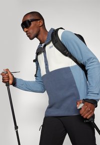 Pullover in pile di colore azzurro chiaro, blu scuro e bianco con colletto a bottoni, dotato di tasca e indossato con occhiali da sole eleganti e uno zaino.