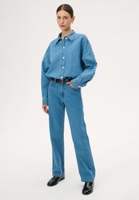 Kobieta stojąca w niebieskiej dżinsowej koszuli zapinanej na guziki, włożonej w wysokowalne niebieskie dżinsy z czarnym paskiem i czarnymi butami na białym tle.