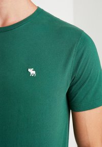 Grünes Baumwoll-T-Shirt mit Rundhalsausschnitt, ausgestattet mit einem kleinen weißen Elch-Logo auf der linken Brust. Glatte Textur, kurze Ärmel.