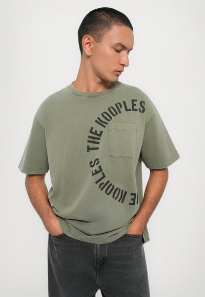 T-shirt de algodão verde oliva com gola redonda, mangas curtas e um bolso frontal. Apresenta texto curvado em preto "THE KOOPLES" na parte da frente.