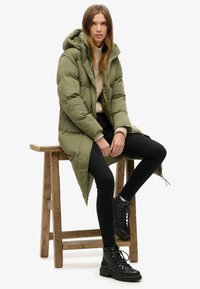 Abrigo de invierno verde acolchado con capucha, suéter de punto texturizado debajo, jeans ajustados negros y botas de tobillo negras con cordones.
