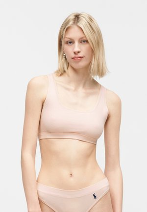 Ξανθιά γυναίκα που φοράει ελαφρύ ροζ αόρατο σουτιέν και ταιριαστό εσώρουχο με ένα μικρό σκουρόχρωμο λογότυπο στο αριστερό ισχίο, στέκεται μπροστά σε απλό φόντο.