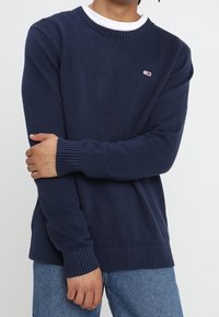 Granatowy sweter dziergany z okrągłym dekoltem i prążkowanymi mankietami. Małe logo na piersi. W zestawie z jasnoniebieskimi dżinsami.