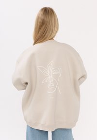 Beżowy, oversize'owy sweatshirt z minimalistycznym białym haftowanym wzorem twarzy i liścia na plecach. Miękka tkanina, ściągacze na rękawach i u dołu.